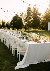Elegant outdoor dinner setup with rustic wooden chairs, a long white table adorned with greenery, grapes, and flowers, under string lights at sunset.