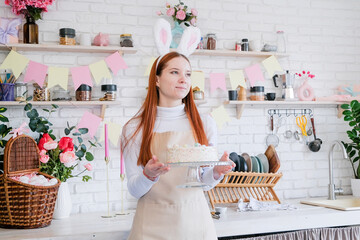 portrait of young woman in bunny ears holding Easter cake in pastel colors in the kitchen