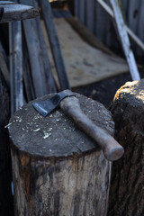 An old ax with a wooden handle lying on a stump used as a block for chopping wood. The background of the photo consists of a rustic yard with scattered logs, old planks and rough wood siding, creating