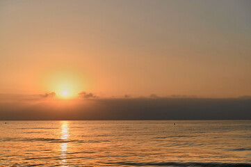 As the sun sets on the horizon, golden hues spread across the sky, illuminating tranquil ocean waters. A few clouds linger above, enhancing the peaceful ambiance of dusk.