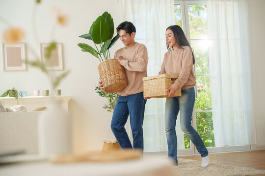 Asian couple carrying woven baskets with a plant and household items, walking through a living space, preparing to settle in their new home. Moving house concept