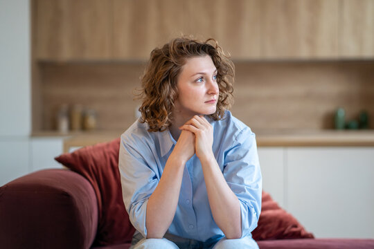 Lonely, distressed woman sitting on sofa at home, lost in deep thoughts. Overwhelmed upset female by anxiety, stress, self-doubt, emotional exhaustion or inner struggles. Uncertainty, low self-esteem.