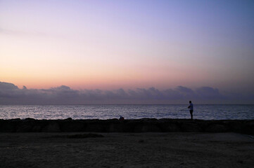 Fototapeta premium Bathed in soft hues of dusk, a person stands contemplatively by the water's edge, watching as the sun sets on the horizon, creating a tranquil atmosphere.
