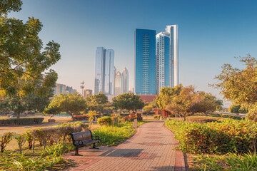 Empty park with bench and brick path leading to modern skyscrapers in Abu Dhabi during sunny morning