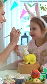 Mother and daughter paint a ceramic bunny together for Easter. The girl, wearing bunny ears, carefully applies yellow paint while her mother assists. The table is filled with colorful paints