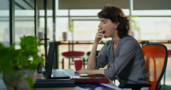Young adult gen z woman lady sitting use laptop do over work time look screen feel bad lazy tired lack dull job office place busy sleepy teen female bored yawning on desk table need break rest