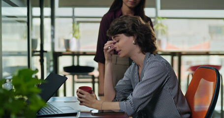 Young adult gen z lazy woman lady sitting do over time work feel bad lack of sleep tired fall asleep on table job office work place bored female girl staff wake up hold tea mug hot coffee cup day