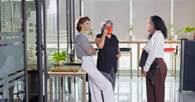 Indian young adult women intern old aged lady hold cup mug drink coffee talk gossip work place expert teen girl staff group team speak idea blab share enjoy fun joy tea break day spend time job office