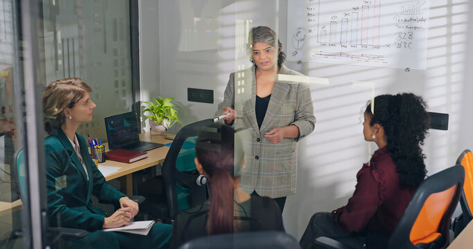 Indian young adult female old elder older boss lady team leader show growth chart white board learn graph data work place teen girl intern expert group talk share stock trade speak idea job office day