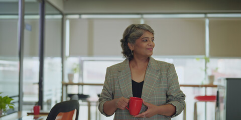 Indian happy old aged boss ceo woman lady hold cup drink coffee routine tea spend break time standing in hall room space smiling elder older relax agent worker staff enjoy day office job work place