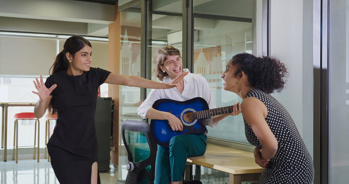 Indian Asian happy young adult gen z girl hold guitar play music make fun joy job office place crazy boss lady teen female multi staff group team do funny dance sing song spend time enjoy work break