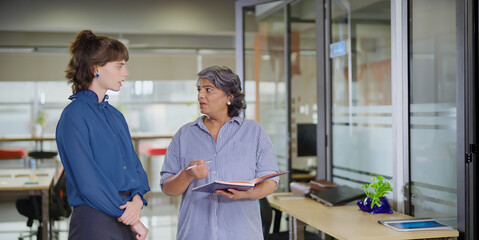 Indian senior old elder boss women lady standing hold book file make new project indoor office young adult expert intern girl older staff do work talk day task read report share ideas plan job place