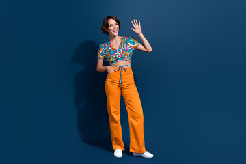 Fototapeta premium Full body photo of pretty young girl waving hand hello greeting dressed stylish flared pants outfit isolated on dark blue color background