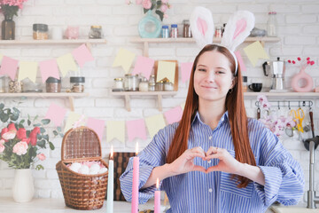 Caucasian woman in bunny ears standing by serving table for Easter dinner, making heart shape with hands