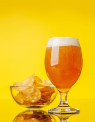 A glass of cold beer alongside a pile of crispy potato chips on a vibrant yellow background