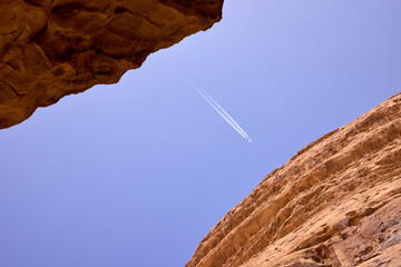 Airplane contrails in blue sky framed by sandstone canyon walls in Jordan. Natural rock formation creating dramatic frame. Adventure travel concept