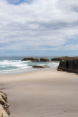 Scenic coastal beach with golden sand, rocky formations, and gentle ocean waves under a partly cloudy sky. A serene seaside landscape