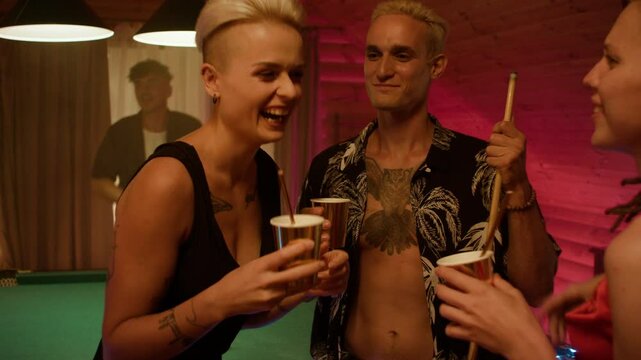 Three friends stand by a billiard table with paper cups. A woman in a black dress laughs while talking with a red-dressed friend with dreads, as a man in a tropical shirt smiles in the background.