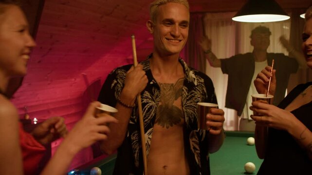 Three friends stand by a billiard table with paper cups. A man in a print shirt holds a cue and cup while a woman in a black dress sips from a straw. In the background, a man dances by curtains.