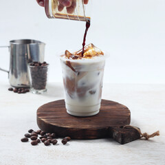 Iced coffee latte, A hand pours dark coffee into a clear glass filled with ice and milk, creating a creamy beverage on a wooden board