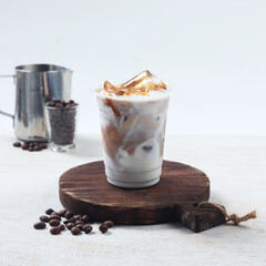 Iced coffee in a clear cup filled with milk and syrup sits on a wooden board, complemented by coffee beans and a metal pitcher