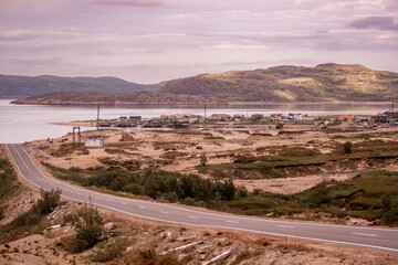 A winding road leads toward a peaceful area by the water in Teriberka, surrounded by hills and sparse vegetation, capturing the tranquility of a sunset over the landscape. Travel in Russia.