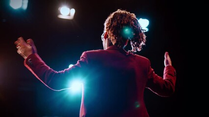 Speaker or host on stage of concert hall, back view against bright soffits. Rearview of charismatic african man with microphone standing alone in front of audience and speaking, motivating life coach