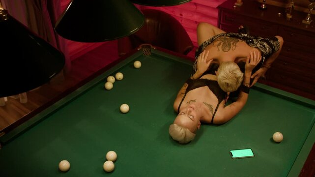 A girl with short hair lies on a billiard table as a man in a floral shirt, barely dressed, kisses her stomach. She holds his shoulders as they draw near, with balls scattered and a phone lit nearby.
