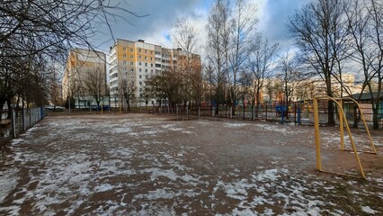 Behind a metal fence surrounded by trees is a stadium with a sandy mini soccer field with goals. Buildings stand nearby. The snow is melting and turning into puddles. The winter sun is breaking © Balser