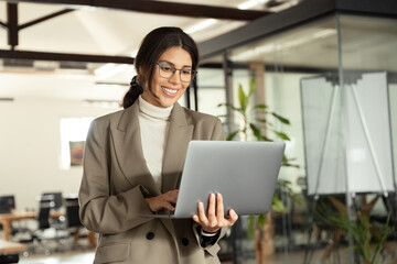Smiling young businesswoman manager holding laptop pc using it fintech application standing at...