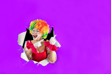 Funny female clown looking through the ripped paper hole pointing at blank template copy spase for advertising, posters, greetings, invitations. Entertainer as Joker in a suit and wig