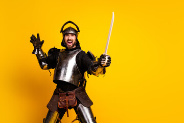 Fototapeta premium Energetic man dressed as a knight posing with a sword in front of a yellow background, emphasizing themes of medieval history
