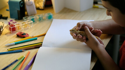 A child holds a leopard gecko while surrounded by colorful pencils and a blank sketchbook on a desk. This photo reflects creativity, curiosity, and a bond with exotic pets.