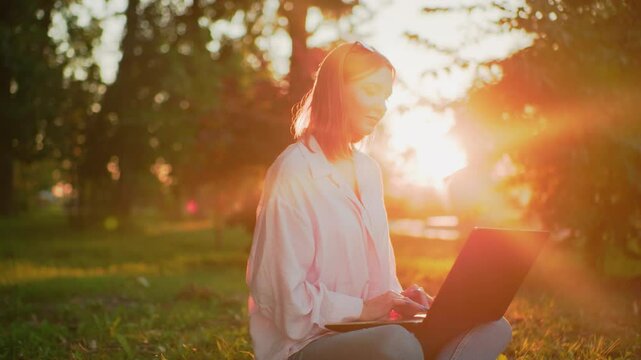 Lady in pink top with glasses resting on her head smiles warmly while typing on laptop, she looks up thoughtfully, then refocuses on her laptop, with sunlight creating a beautiful glow around her