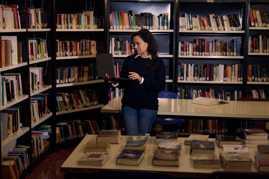 Woman Using Laptop in Library Surrounded by Bookshelves and Tables