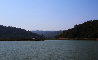 The water in the lake curves along the mountain. With a canopy and flag visible under the shade of trees. The sky is clear.

