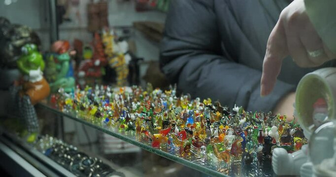 Retail trade. The hands of a woman who chooses to buy small figurines and souvenirs made of colored glass in large numbers in the window of a small store.