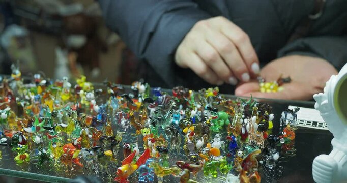 Retail trade. The hands of a woman who chooses to buy small figurines and souvenirs made of colored glass in large numbers in the window of a small store.