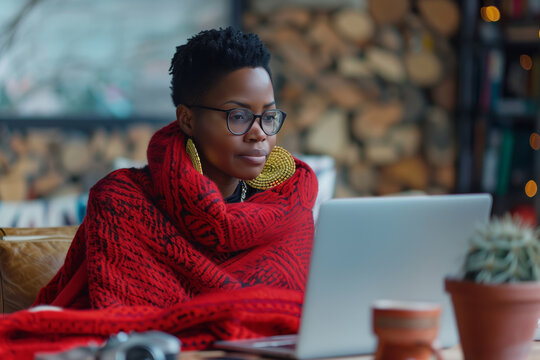African woman wearing glasses working at home. Concept of comfort and relaxation, as the woman is wrapped in a warm blanket while working on her laptop