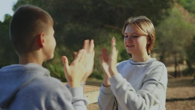 Teenage friends sharing joyful moment at sunny park, playing rhythmic hand-clapping game while sitting on picnic table, expressing carefree childhood connection and happiness
