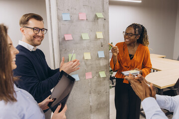 Collaborative team analyzing project strategy, placing multicolored sticky notes on glass wall in bright modern office setting