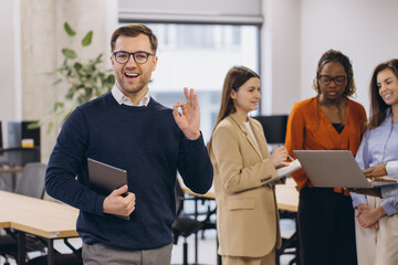 Smiling businessman showing ok sign holding tablet with diverse colleagues working on laptop in modern office, teamwork and success concept