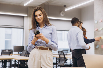 Businesswoman using mobile phone in modern office while colleagues working together on project in...