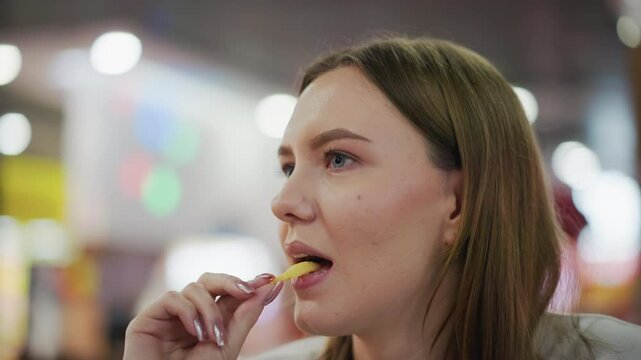 Close-up of lady eating fries with a focused look, soft bokeh background with colorful blinking lights in a lively shopping mall setting