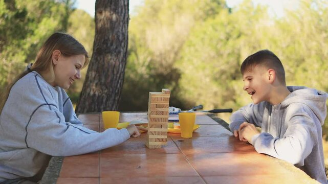 Young friends balancing wooden jenga blocks with concentrated play, sharing outdoor summer fun amid verdant forest greenery and mutual excitement.Happy friendship day