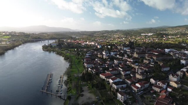 Descending aerial view of Tui, featuring its historic cathedral overlooking the calm Mi&ntilde;o River, the natural border between Spain and Portugal. Tui-Valen&ccedil;a International Bridge and marina are visible.