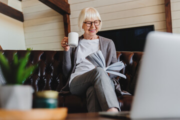 Old retired 70s lady reading book at home, drinking hot tea, resting on sofa in cozy living room. Mature reader relaxing with novel over cup of coffee or chocolate.