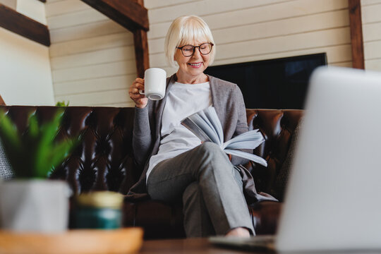 Old retired 70s lady reading book at home, drinking hot tea, resting on sofa in cozy living room. Mature reader relaxing with novel over cup of coffee or chocolate.