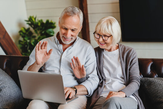 Aged couple sitting on couch in living room country house make videocall look at pc screen wave hands greeting friend, grown up children, grandchildren, modern tech and older generation users concept