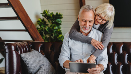 Couple of seniors looking at the same tablet hugged on the sofa indoor at home. Caucasians mature retired man and woman using technology sitting on couch in living room. Older generation users concept